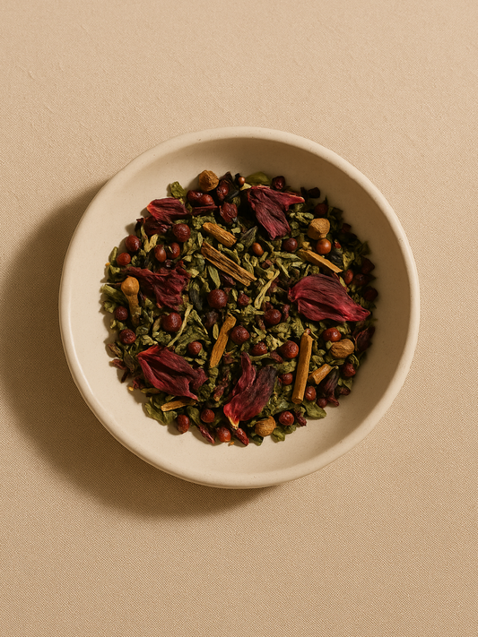 Overhead view of loose Flow Tea herbs in a ceramic bowl, showing hibiscus petals, cinnamon bark, and schisandra berries and nettle leaves on a soft neutral background.